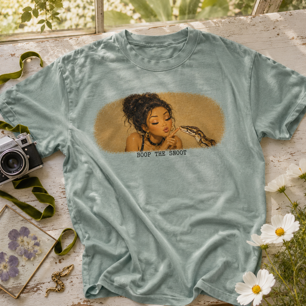 A light blue-green graphic t-shirt laid flat on a rustic white wooden surface, surrounded by vintage props including a camera, green ribbon, pressed flowers, and white daisies. The shirt features a central graphic of a woman with dark hair in a vintage illustration style, with the text 'BOOP THE SMOOT' printed below.