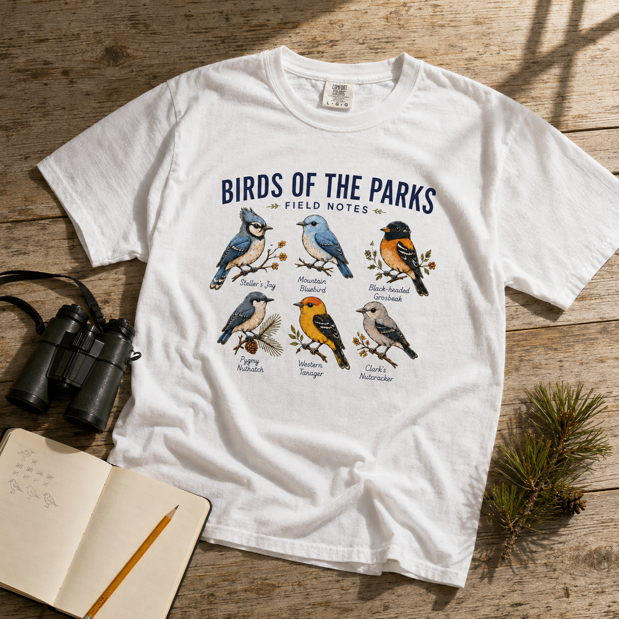 Birds of the Parks Field Notes white Comfort Colors tee styled with binoculars and a field notebook in strong sunlight